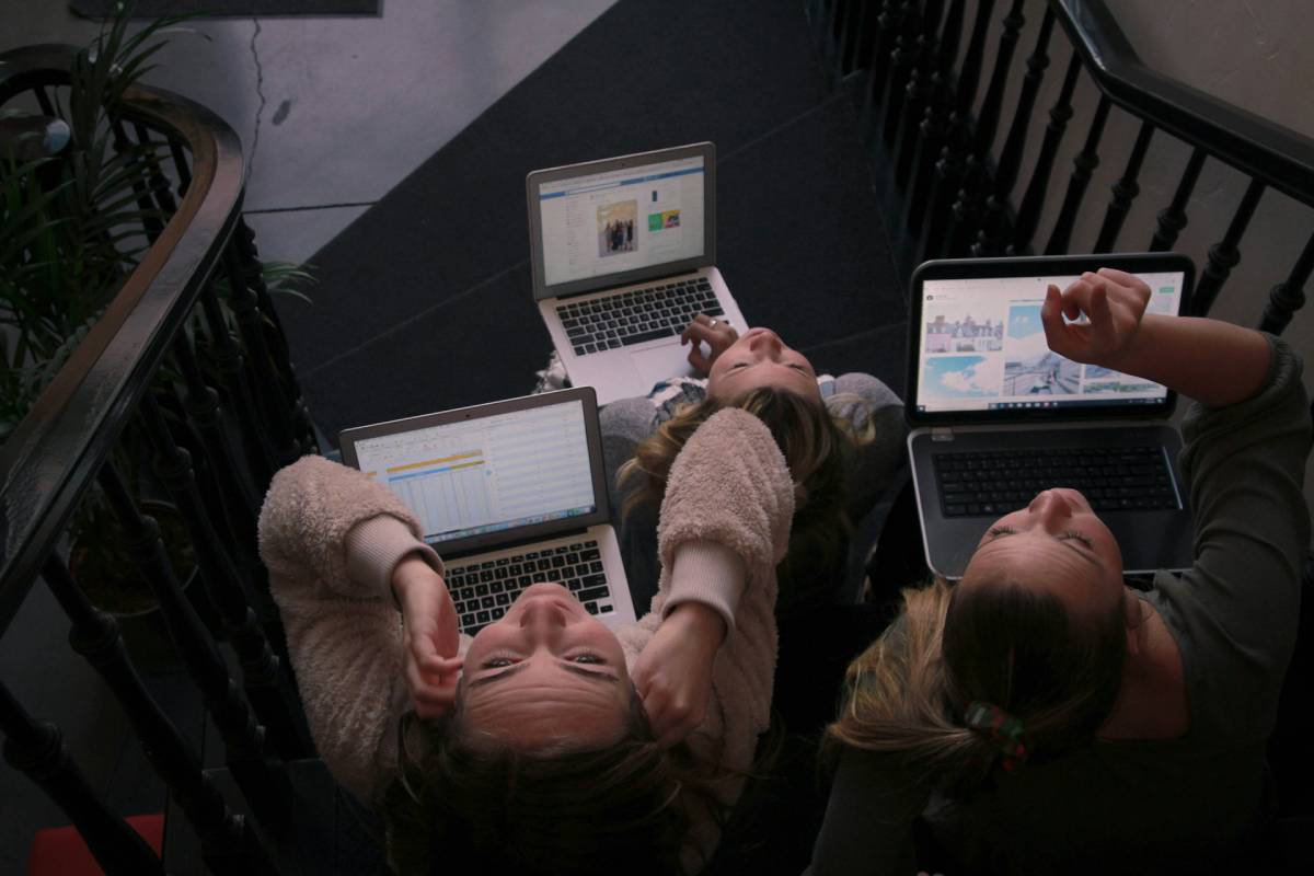 na zdjęciu trzy kobiety w różnym wieku siedzą stłoczone na schodach z laptopami na kolanach