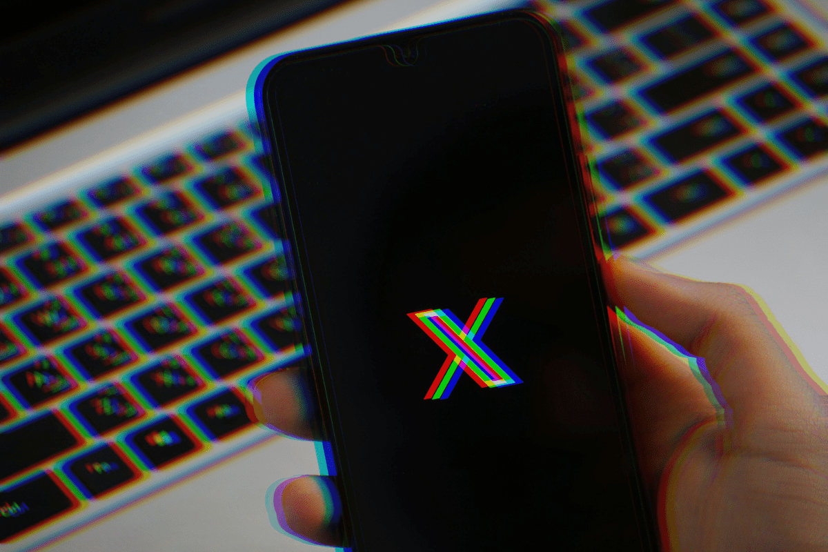 na rozmytym zdjęciu dłoń trzymająca smartfona z wyświetlonym logo X